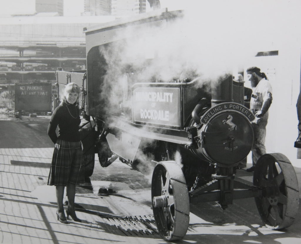 A young woman wearing a kilt is looking at the photographer and stands next to a steam wagon. The steam wagon, which is surrounded by clouds of steam, is standing on a concrete pavement outside a workshop. A steam wagon is a self-propelled steam-powered truck with a timber tip tray. The side of the steam wagon has the wording 'Municipality of Rockdale' for whom the wagon was built and the front has the manufacturer's name, 'Aveling & Porter'. A man with a long beard is standing on the other side of the wagon looking at the wagon's steam engine. 